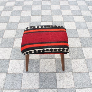 May include: A small, square stool with wooden legs and a red, black, and white striped cushion. The cushion has a black and white pom-pom trim.