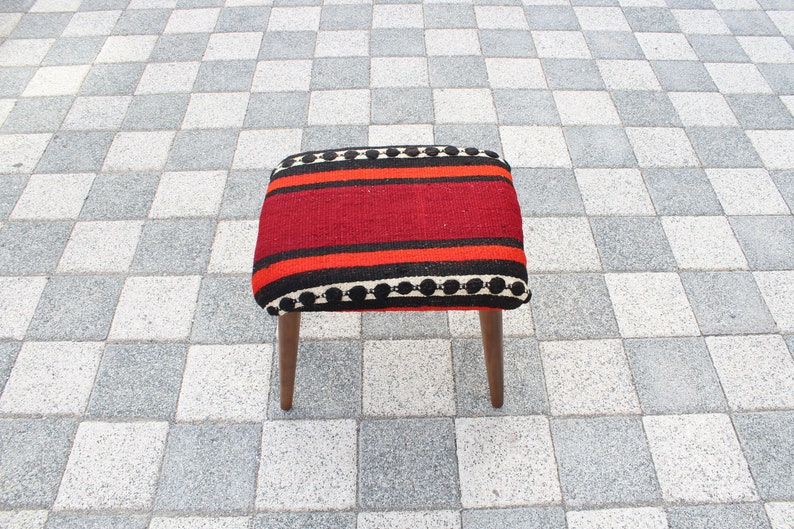 May include: A small, square stool with wooden legs and a red, black, and white striped cushion. The cushion has a black and white pom-pom trim.