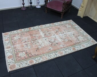 May include: A rectangular, distressed-look rug with a peach and cream color palette. The rug features a central floral pattern with a cream border and a decorative edge. The rug is placed on a black tiled floor.