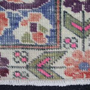 May include: Close-up of a beige rug with a fringe border. The rug has a geometric pattern with shades of blue, red, green, and purple.