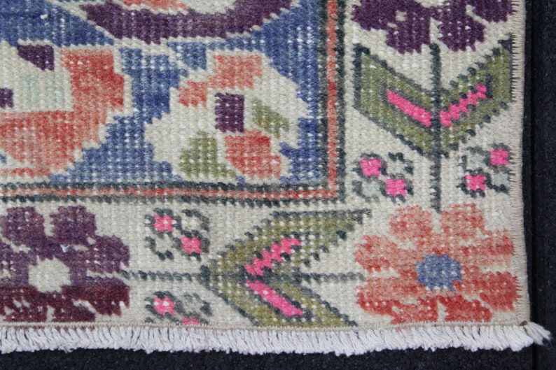 May include: Close-up of a beige rug with a fringe border. The rug has a geometric pattern with shades of blue, red, green, and purple.