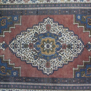 May include: A vintage rug with a red background and a central medallion featuring a blue, yellow, and green floral design. The rug has a border with a repeating pattern of blue, white, and red.