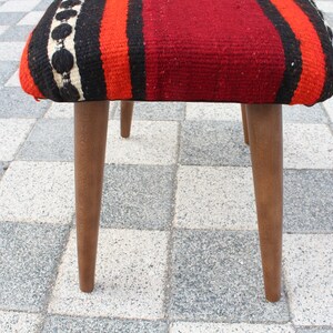 May include: A wooden stool with a red, orange, black, and white striped fabric seat. The stool has three legs and is sitting on a checkered floor.