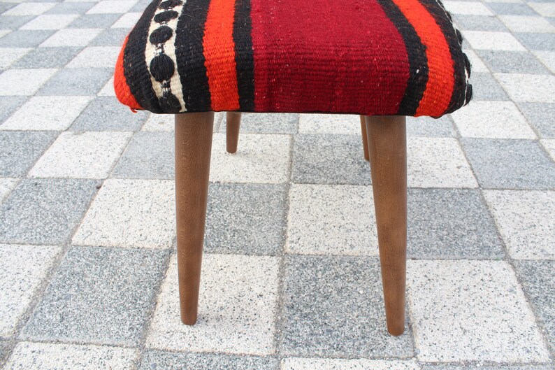 May include: A wooden stool with a red, orange, black, and white striped fabric seat. The stool has three legs and is sitting on a checkered floor.