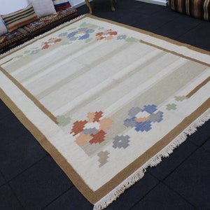 Puede incluir: Una alfombra de color crema con un borde marr&oacute;n y dise&ntilde;os florales en tonos de azul, naranja y verde. La alfombra tiene un borde de flecos.