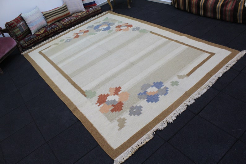 Puede incluir: Una alfombra de color crema con un borde marr&oacute;n y dise&ntilde;os florales en tonos de azul, naranja y verde. La alfombra tiene un borde de flecos.