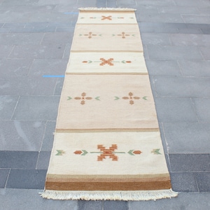 May include: A beige runner rug with a geometric pattern in shades of brown and green. The rug has a fringe border.