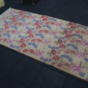 May include: A rectangular rug with a beige background and a floral pattern in red, blue, and pink. The rug has a distressed look, with faded colors and a worn appearance. The edges are finished with a white fringe.
