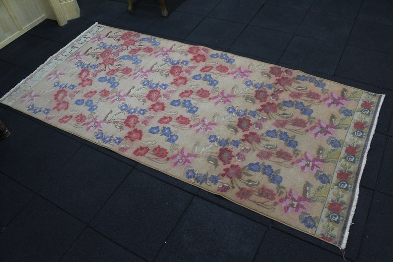 May include: A rectangular rug with a beige background and a floral pattern in red, blue, and pink. The rug has a distressed look, with faded colors and a worn appearance. The edges are finished with a white fringe.