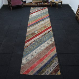 May include: A long, rectangular, multi-colored rug with a striped pattern. The rug is made of different fabrics and has a vintage look. The rug is laid out on a black tiled floor.