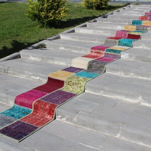 May include: A colourful patchwork runner rug made from different pieces of fabric in shades of red, pink, purple, blue, green, and yellow. The rug is laid out on a set of concrete steps.