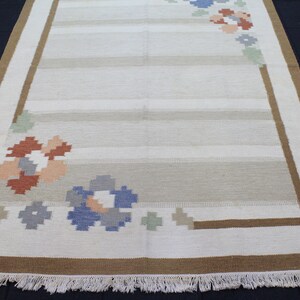 Puede incluir: Una alfombra blanca con un borde marr&oacute;n y un dise&ntilde;o floral en tonos azules, rosas y naranjas. La alfombra tiene un borde de flecos.