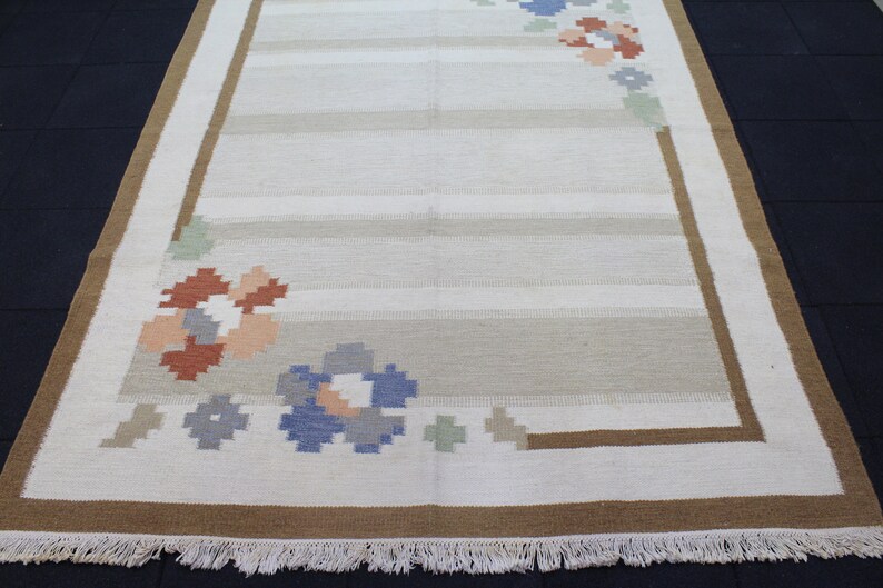 Puede incluir: Una alfombra blanca con un borde marr&oacute;n y un dise&ntilde;o floral en tonos azules, rosas y naranjas. La alfombra tiene un borde de flecos.