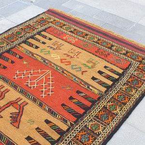 May include: A traditional rug with a geometric pattern in shades of red, brown, black, and green. The rug has a fringe border and is made of wool.