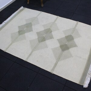 May include: A white rug with a geometric pattern of green diamonds. The rug has fringe on all sides.