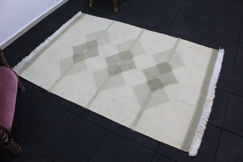 May include: A white rug with a geometric pattern of green diamonds. The rug has fringe on all sides.