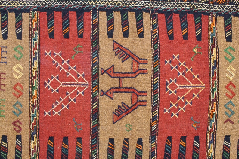 May include: A close-up of a colorful woven rug with geometric patterns and stylized animal figures. The rug features a red, brown, and beige color scheme with accents of green, blue, and black.