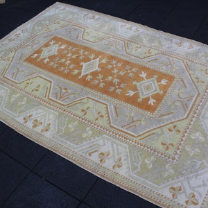 May include: A vintage Turkish rug with a beige background and a central orange rectangle with a geometric pattern. The rug has a white border with a repeating pattern of stylized flowers.