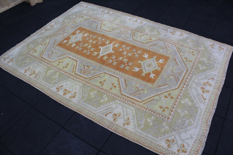 May include: A vintage Turkish rug with a beige background and a central orange rectangle with a geometric pattern. The rug has a white border with a repeating pattern of stylized flowers.