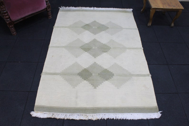 May include: A white rug with a geometric pattern of light green diamonds. The rug has a fringe border.