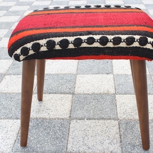May include: A red, black, and white striped ottoman with a wooden frame and legs. The ottoman has a woven pattern with black circles.