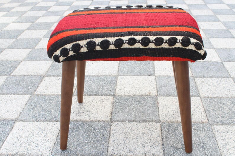May include: A red, black, and white striped ottoman with a wooden frame and legs. The ottoman has a woven pattern with black circles.