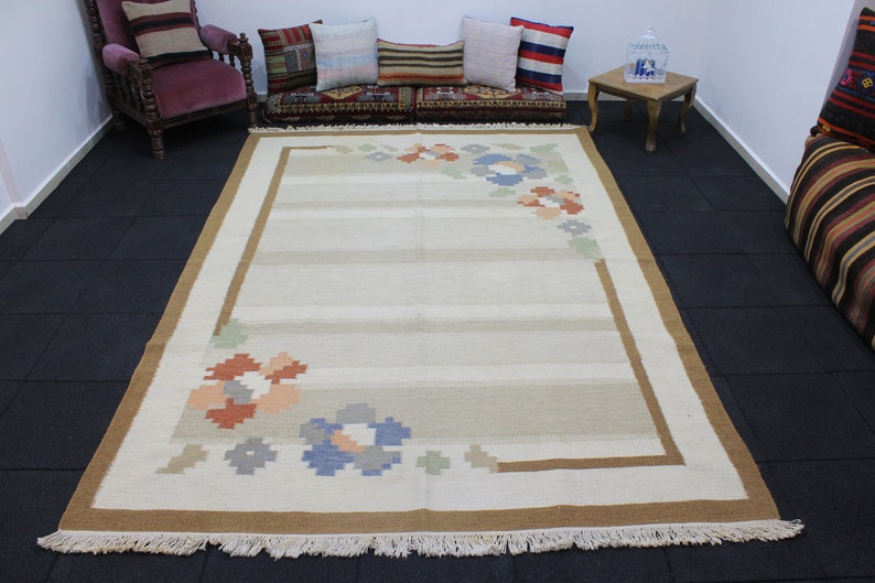 Puede incluir: Una alfombra tejida a mano de color blanco y beige con un borde marr&oacute;n y dise&ntilde;os florales en las esquinas. La alfombra tiene flecos alrededor de los bordes.