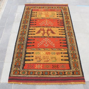 May include: A traditional Turkish rug with a red, orange, and black geometric pattern. The rug has a fringe border and is made of wool.