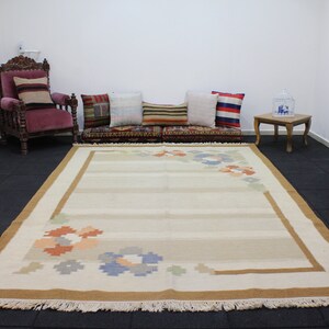 Puede incluir: Una alfombra beige y marr&oacute;n con un dise&ntilde;o floral. La alfombra tiene un borde de flecos y est&aacute; colocada sobre un suelo de baldosas negras.