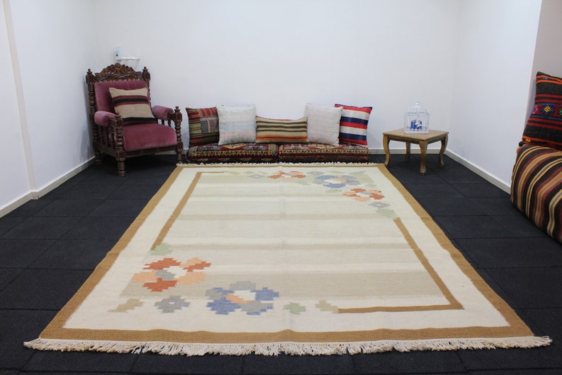 Puede incluir: Una alfombra beige y marr&oacute;n con un dise&ntilde;o floral. La alfombra tiene un borde de flecos y est&aacute; colocada sobre un suelo de baldosas negras.