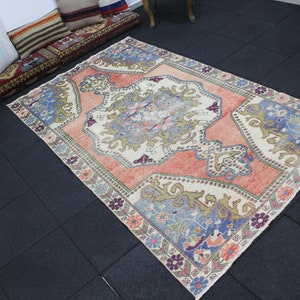May include: A vintage Turkish rug with a cream background and a large, intricate floral design in shades of pink, blue, and green. The rug has a faded, distressed look, giving it a unique and antique feel.
