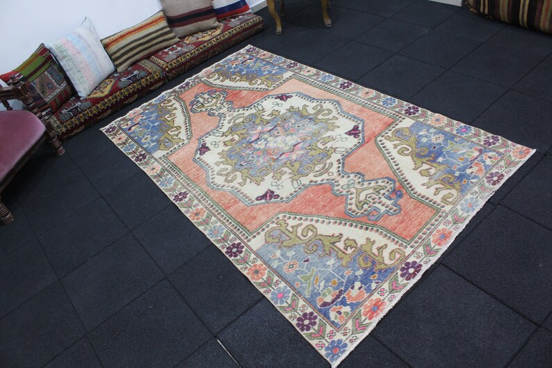 May include: A vintage Turkish rug with a cream background and a large, intricate floral design in shades of pink, blue, and green. The rug has a faded, distressed look, giving it a unique and antique feel.