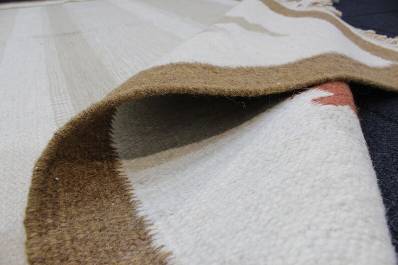 Puede incluir: Primer plano de una alfombra tejida blanca y marr&oacute;n con una superficie texturizada. La alfombra tiene un borde marr&oacute;n y un centro blanco con un patr&oacute;n sutil.