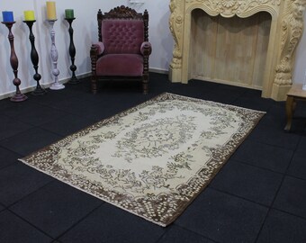 May include: A cream-colored rug with a brown floral pattern. The rug is laid out on a black tiled floor in a room with a wooden fireplace and a pink armchair.