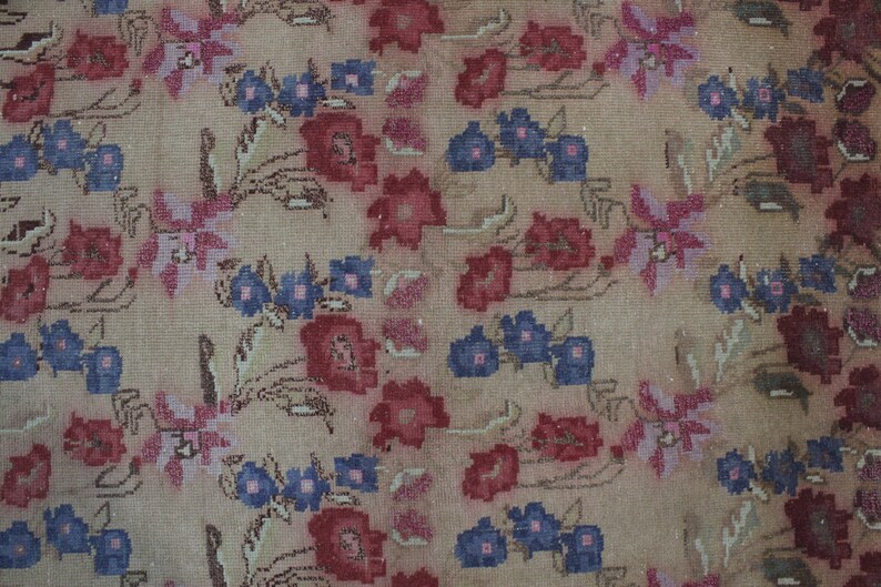 May include: A beige floral rug with a repeating pattern of red, pink, and blue flowers. The rug has a distressed look, with faded colors and a worn texture. The flowers are stylized and arranged in a symmetrical design.