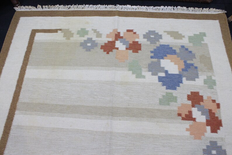 Puede incluir: Una alfombra blanca con un borde marr&oacute;n y un dise&ntilde;o floral en tonos de azul, verde, rojo y rosa.