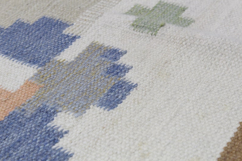 Puede incluir: Primer plano de una alfombra tejida con un patr&oacute;n geom&eacute;trico en tonos azules, verdes y beige. La alfombra tiene un fondo blanco y una superficie texturizada.