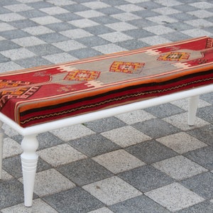 May include: White wooden bench with a red, orange and beige patterned cushion. The cushion is made of woven fabric and has a traditional design.