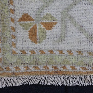 May include: Close-up of the bottom edge of a beige rug with a geometric pattern in shades of brown and green. The rug has a fringe border.