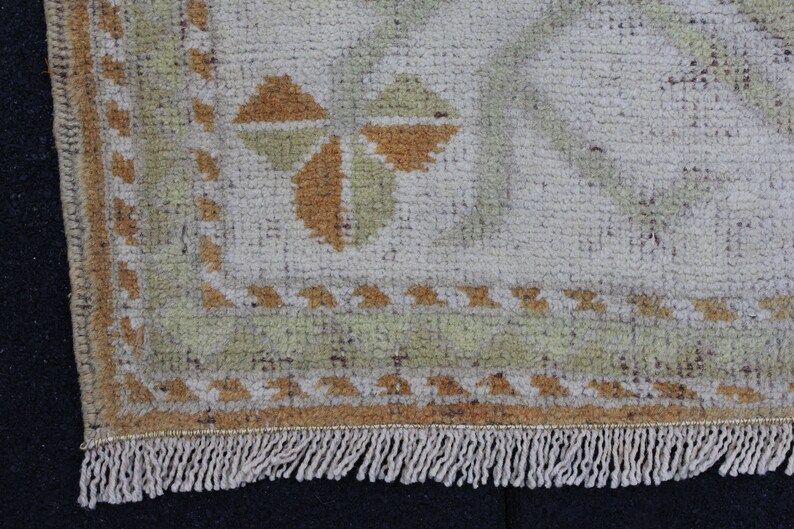 May include: Close-up of the bottom edge of a beige rug with a geometric pattern in shades of brown and green. The rug has a fringe border.