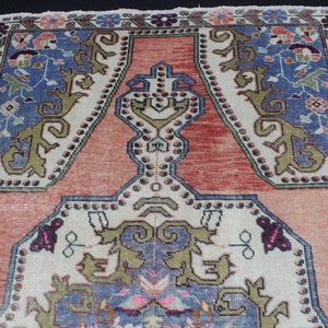 May include: A vintage Turkish rug with a traditional design. The rug has a cream background with a red center and blue and green accents. The rug is decorated with floral and geometric patterns.