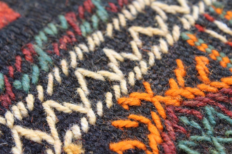 May include: Close-up of a woven fabric with a geometric pattern. The fabric is black with white, orange, red, and green threads.