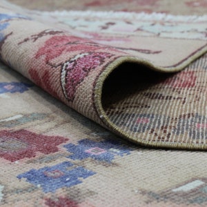 May include: Beige rug with a floral pattern in shades of red, pink, and blue. The rug is rolled up, revealing the underside with a striped pattern. The rug's edges are finished with a brown border.