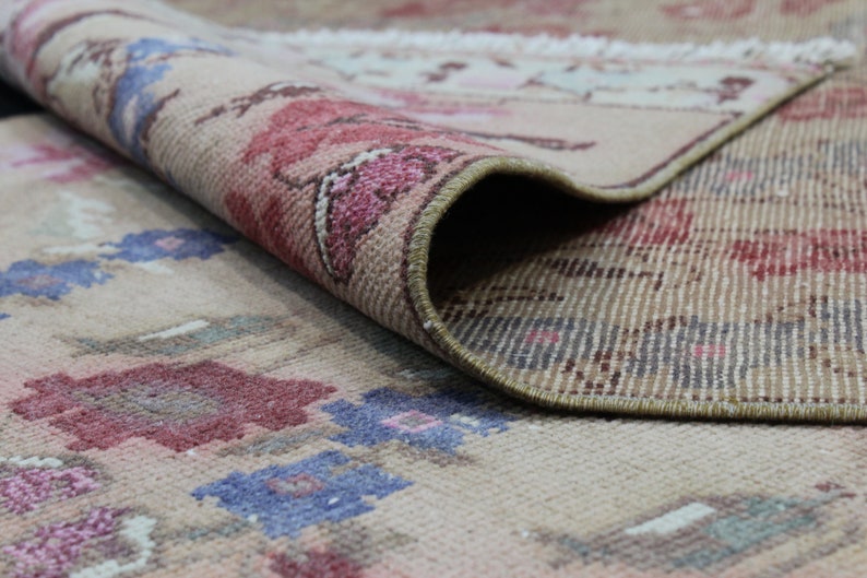 May include: Beige rug with a floral pattern in shades of red, pink, and blue. The rug is rolled up, revealing the underside with a striped pattern. The rug's edges are finished with a brown border.