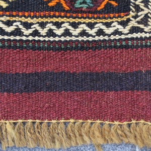 May include: A close-up of a woven rug with a burgundy, black, and white striped pattern. The rug has a fringe border made of natural fibers.