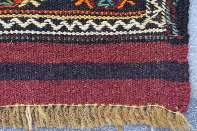 May include: A close-up of a woven rug with a burgundy, black, and white striped pattern. The rug has a fringe border made of natural fibers.