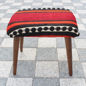 May include: A red, black, and white striped ottoman with wooden legs. The ottoman has a woven fabric top with a black and white trim.