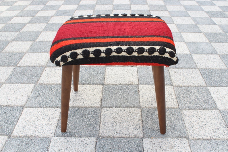 May include: A red, black, and white striped ottoman with wooden legs. The ottoman has a woven fabric top with a black and white trim.