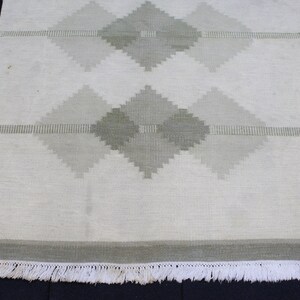 May include: A white woven rug with a geometric pattern of green diamonds. The rug has a white fringe border.
