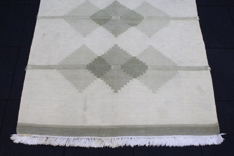 May include: A white woven rug with a geometric pattern of green diamonds. The rug has a white fringe border.